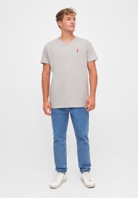 Young man standing wearing a light gray V-neck t-shirt, blue jeans, and white sneakers against a plain white background.
