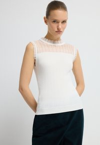 Ærmeløs hvid top med ribbet design, der har et gennemsigtigt stribet panel ved halsudskæringen og bølgede kanter. Kombineret med mørkegrønne bukser.