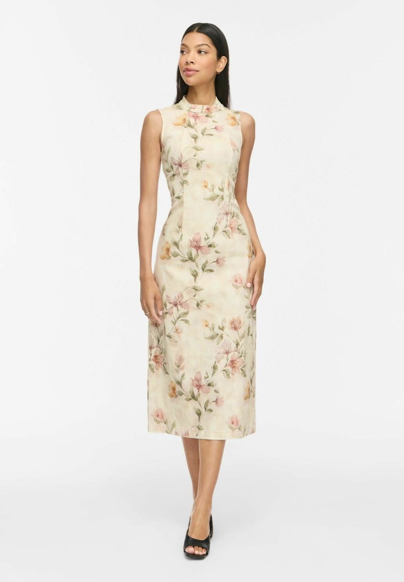 Femme portant une robe crème sans manches, longueur genou, avec un col haut et un motif floral rose et vert, debout, les mains détendues le long du corps.