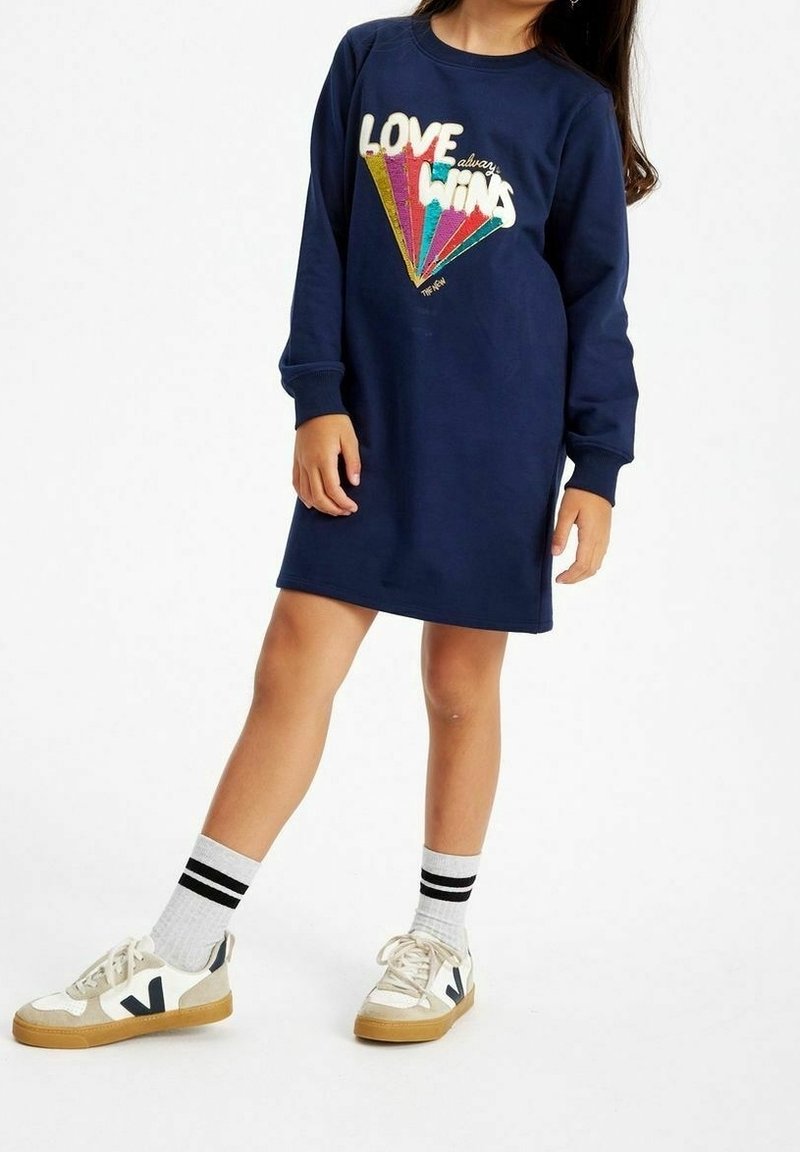 Granatowa sukienka-sweterek z długimi rękawami, z wielokolorowym napisem "MIŁOŚĆ zawsze WYGRYWA", w połączeniu z białymi trampkami i w paski skarpetkami.