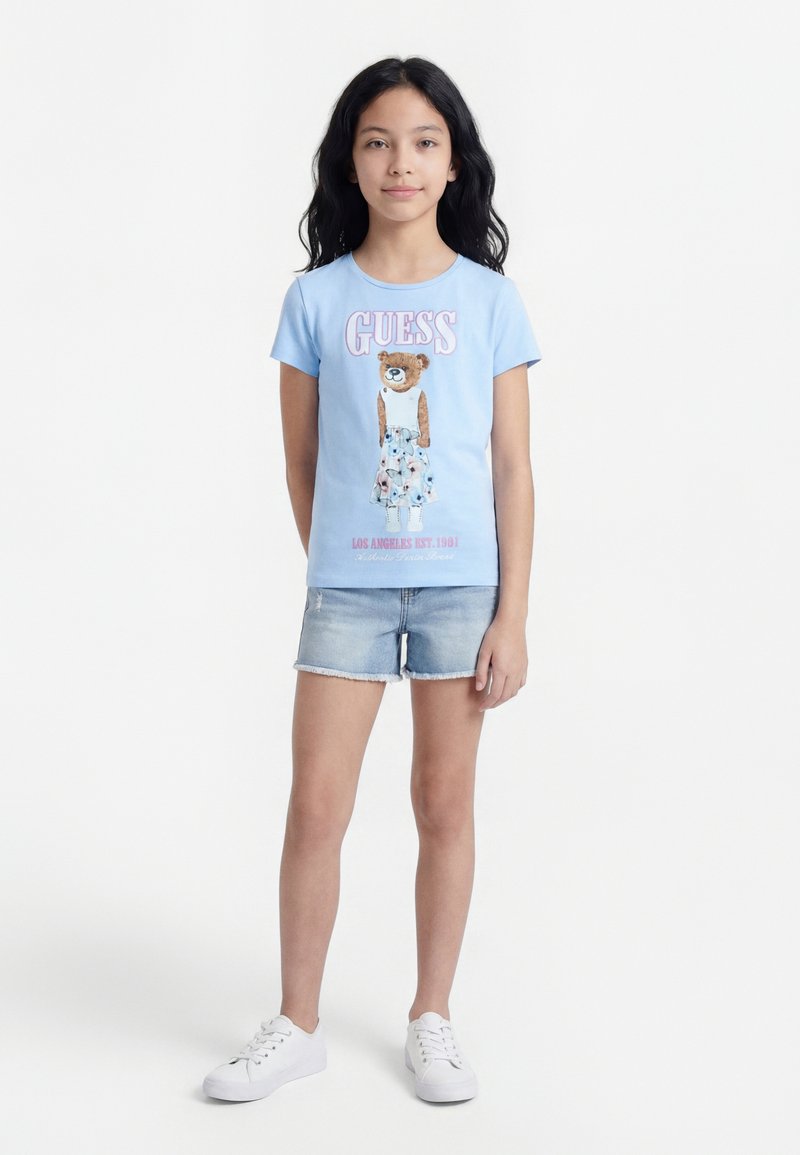 Νεαρή κοπέλα με μακριά σκούρα μαλλιά, ντυμένη με ανοιχτό γαλάζιο T-shirt με γραφικό αρκουδάκι, τζιν σορτς και λευκά αθλητικά παπούτσια, στέκεται μπροστά σε ένα απλό λευκό φόντο.