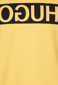 Camiseta amarilla de manga corta con una audaz banda horizontal negra con el texto "HUGO" en el pecho. Tela suave, ajuste casual.