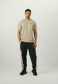 Bežové tričko s krátkym rukávom a logom. Čierne nohavice majú červené a šedé bočné pruhy. Model má na sebe čierne tenisky a stojí čelom.