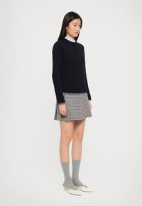 Námornícky sveter s rebrovaným lemom, skombinovaný s kockovanou sukňou. Šedé ponožky a biele špicaté topánky dopĺňajú outfit. Minimalistický dizajn.