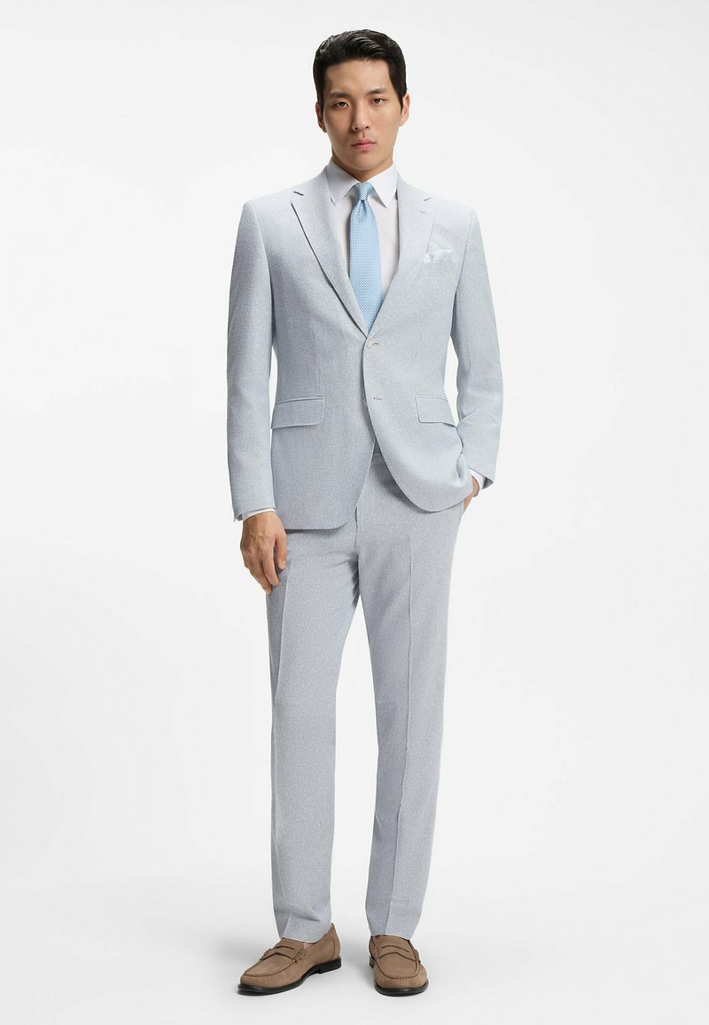 Homme en costume gris clair avec chemise blanche, cravate bleu clair, mocassins marron, debout avec une main dans la poche devant un fond blanc.