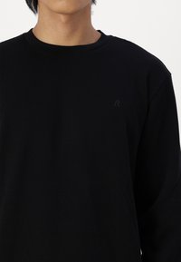 Maglietta nera a maniche lunghe in tessuto rigato con scollo rotondo e un sottile logo tono su tono sulla parte superiore sinistra. Realizzata in tessuto testurizzato.