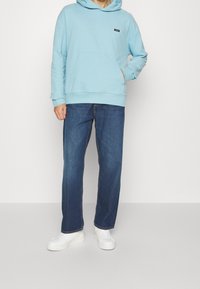 Felpa leggera blu con tasca frontale e un piccolo logo, abbinata a jeans dritti blu scuro e sneaker bianche.