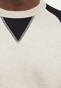 Κοντινή λήψη μπεζ και μαύρου φούτερ με στρογγυλή λαιμόκοψη που δείχνει λεπτομέρειες ραφής και πάνελ υφάσματος με αντίθεση κάτω από το γιακά.