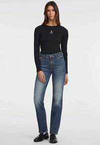 Γυναίκα με μαύρο μπλουζάκι μακρύ μανίκι με μικρό λογότυπο, μπλε ψηλόμεσα τζιν και μαύρες μοκέτες, που στέκεται μπροστά σε απλή φόντο.