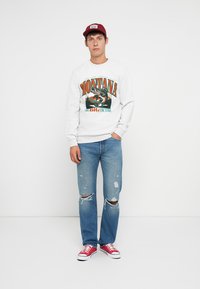 Sweatshirt branco com gráfico "MONTANA", jeans de ganga com características desgastadas e ténis de lona vermelhos. Homem usa um boné de basebol vermelho.