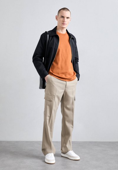 Sort lynlåsjakke over en orange sweater, beige cargo bukser og hvide sneakers. Glatte teksturer med synlige syninger og lommer.