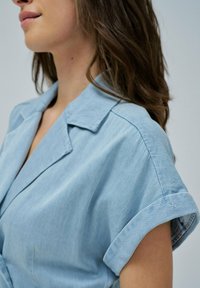 Camisa de mezclilla azul claro con cuello y mangas cortas enrolladas, que presenta un discreto detalle del logo en la manga.