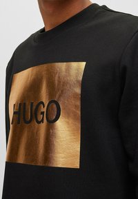 Sweat-shirt noir avec un logo rectangulaire métallique doré portant l'inscription "HUGO" en lettres noires épaisses, col et poignets côtelés.