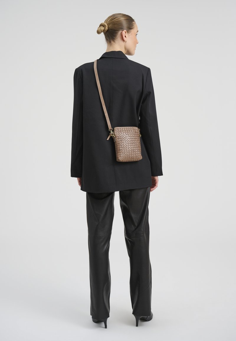 Femme aux cheveux blonds attachés en chignon, portant un long blazer noir, un pantalon en cuir noir et un sac bandoulière tressé marron, vue de dos.