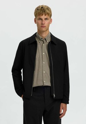 Homme blond portant une chemise beige à carreaux boutonnée sous une veste noire zippée et un pantalon foncé, debout devant un fond clair uni.