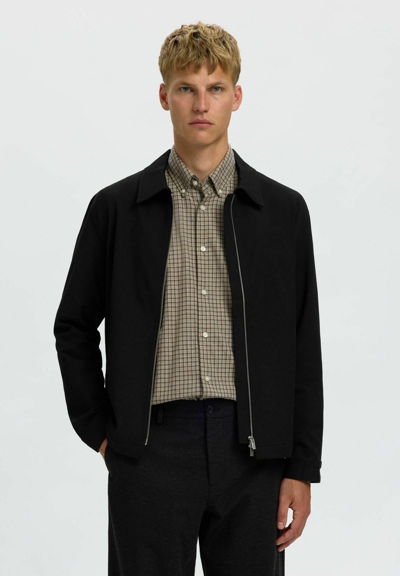 Homme blond portant une chemise beige à carreaux boutonnée sous une veste noire zippée et un pantalon foncé, debout devant un fond clair uni.