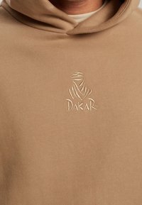 Sudadera con capucha beige con el logo bordado de Dakar que muestra una figura estilizada con un pañuelo en la cabeza, centrada en el pecho.