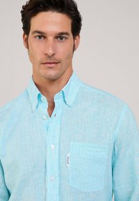 Camisa de botones a rayas en azul claro y blanco, con collar, mangas largas y un bolsillo en el pecho que tiene una etiqueta de la marca. Textura de tela de algodón.