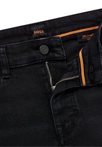 Jeans de mezclilla negra con cremallera plateada parcialmente desabotonada, botón negro grabado con "BOSS" y detalles de costura naranja en el interior de la cinturilla.