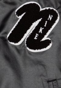 Juoda ir balta Nike logo detalė su puriu tekstūru, apsupta baltos medžiagos, prieš tamsiai pilką lygų foną.