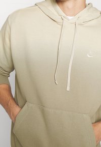 Felpa Nike beige con design a gradient, dotata di tasca anteriore, cappuccio con coulisse e logo ricamato sul petto. Texture morbida.