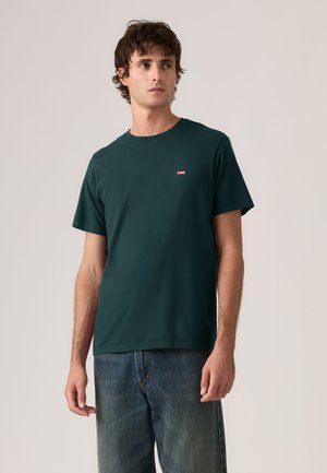 Giovane uomo che indossa una maglietta Levi's verde scuro e jeans blu, in piedi e che guarda leggermente verso sinistra su uno sfondo uniforme.