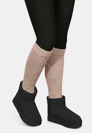 Schwarze Stiefeletten aus strukturiertem Stoff, kombiniert mit hellrosa grob gestrickten Beinwärmern, die sich am oberen Rand sammeln. Schwarze Leggings vervollständigen das Outfit.