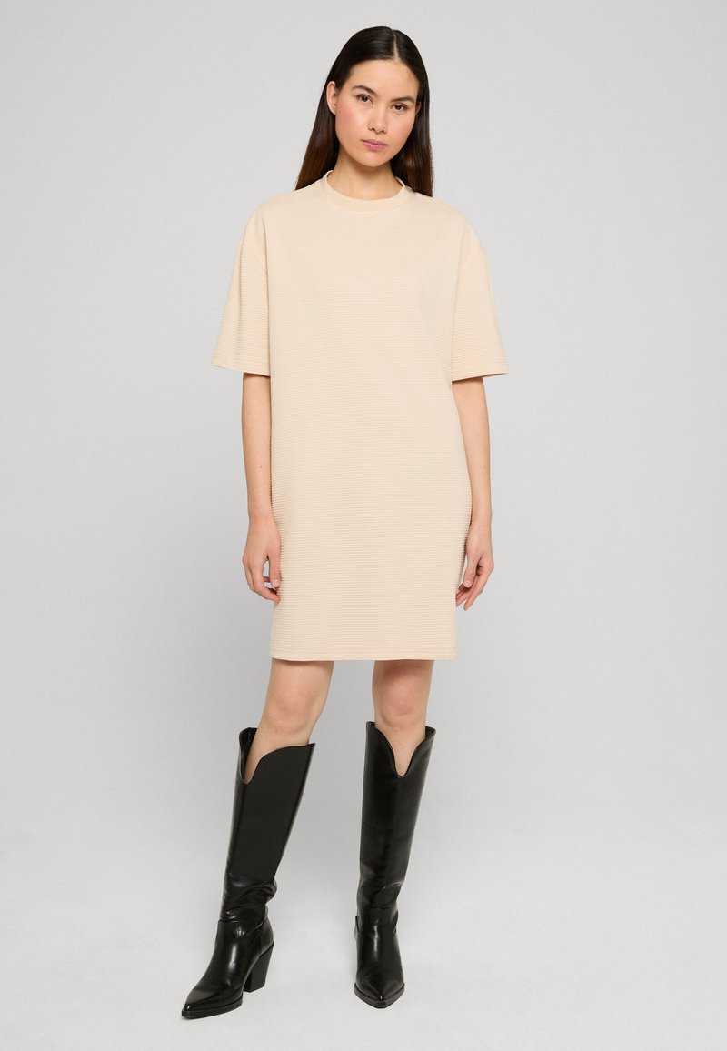 Femme debout portant une robe beige à manches courtes jusqu'au genou et des bottes à talons noires montant jusqu'au genou, sur un fond clair uni.