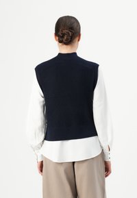 Frau mit einem Haarknoten, die eine marineblaue Strickweste über einer weißen Bluse und beigen weiten Hosen trägt, mit dem Rücken zu einer einfarbigen Hintergrund gerichtet.