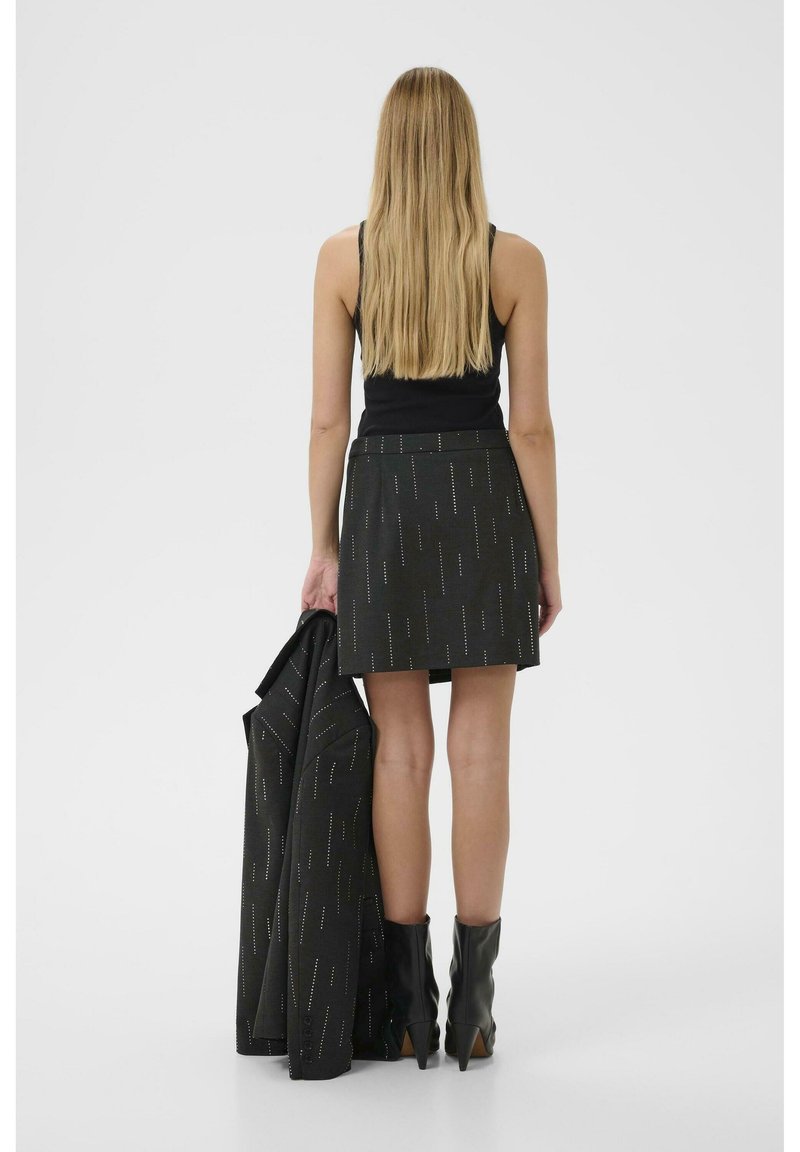 Gonna nera con motivi verticali in filo metallico, abbinata a un blazer coordinato tenuto in una mano. La modella indossa stivali neri alla caviglia.