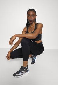 Črne atletske kratke legging hlače v kompletu s črnim športnim modrčkom. Sivi in modri tekaški čevlji s tremi črtami in teksturiranim dizajnom.