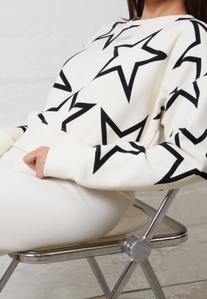 Persoon zittend op een metalen stoel, gekleed in een witte trui met grote zwarte sterpatronen en witte broek.