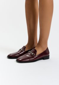 Burgundské kožené mokasíny se stříbrnými kovovými detaily, čtveratou špičkou a plochými černými podrážkami, které vynikají elegantním designem a hladkým povrchem.