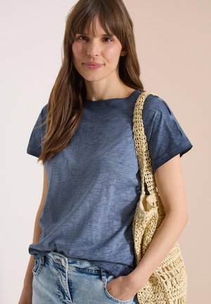 Kvinde med langt brunt hår iført en blå t-shirt og jeans, bærende en beige hæklede skuldertaske, smilende diskret.