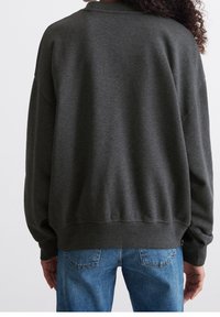 Personne portant un sweat-shirt gris foncé à manches longues et un jean bleu, de dos avec des cheveux bouclés visibles sur le côté droit.