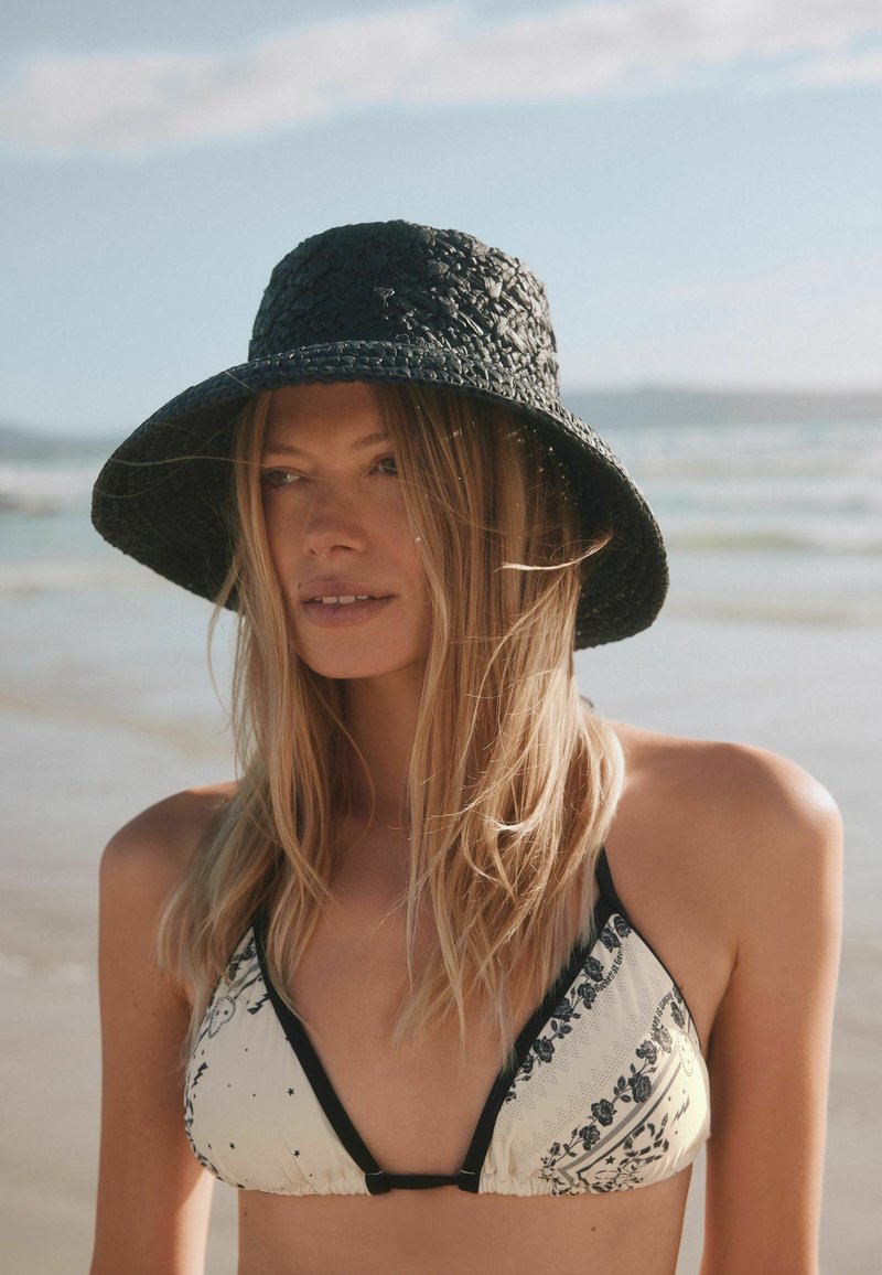 Donna con capelli biondi lunghi che indossa un cappello nero a tesa larga e un top bikini decorato, in piedi su una spiaggia con il mare e il cielo sullo sfondo.