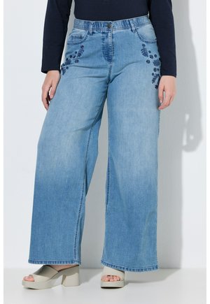 Person trägt hellblaue Jeans mit weitem Bein und dunkelblauer Blumenstickerei an den Hüften, kombiniert mit einem schwarzen Oberteil und beigen Sandalen mit Blockabsatz.