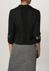 Veste courte noire à manches trois quarts, ornée d'accents brodés sur les poignets et d'une texture lisse semblable à du satin.