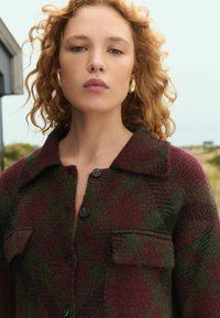 Burgundy and green plaid shirt jacket made of textured fabric, featuring a button front, collar, and chest pockets.