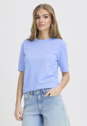 Giovane donna con lunghi capelli biondi, indossa una maglietta celeste a maniche corte e jeans chiari, in piedi con una mano in tasca su uno sfondo semplice.