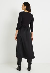 Largo vestido negro con parte superior ajustada y falda de satén fluida, con mangas tres cuartos y cintura ceñida. Sin patrones o detalles visibles.