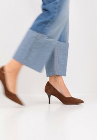 Escarpin à talon haut en daim marron, avec un bout pointu et un talon élancé, porté avec un pantalon en denim à jambes larges présentant des ourlets effilochés et une teinte bleu clair.