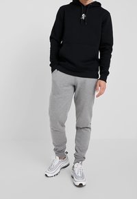 Felpa nera con cappuccio e tasca frontale, abbinata a pantaloni della tuta grigi e sneakers bianche con dettagli grigi. Design minimalista, abbigliamento casual.