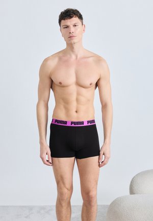 Άνδρας χωρίς πουκάμισο στέκεται μπροστά σε ένα απλό φόντο, φοράει μαύρα μαγιό μπόξερ με μωβ λάστιχο μέσης και λογότυπο Puma.