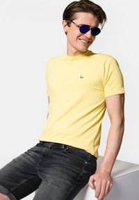 Żółta koszulka z krótkim rękawem i okrągłym dekoltem, z małym zielonym logo na piersi, w zestawieniu z ciemnoszarymi szortami dżinsowymi.