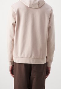 Sudadera con capucha de color beige claro y ajuste relajado, de tela texturizada y con bajo acanalado. Combinada con pantalones marrones, mostrando las mangas ligeramente remangadas.