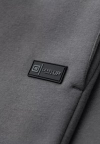Grauer Stoff mit glatter Textur, der ein schwarzes rechteckiges Etikett mit der Prägung "LEVEL UP" und einem runden Logo aufweist.
