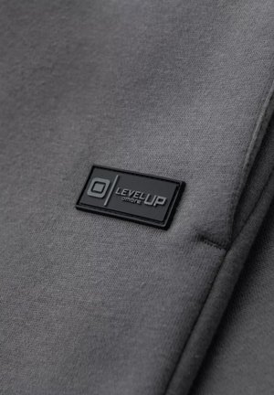 Grå stof med en glat tekstur, der har en sort rektangulær mærkat med præget tekst "LEVEL UP" og et rundt logo.