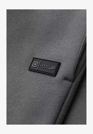 Tissu gris au toucher lisse, avec une étiquette rectangulaire noire embossée portant la mention "LEVEL UP" et un logo circulaire.