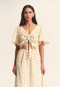 Kremna majica z zlatimi pikami, kratkimi razgibanimi rokavi, V-izrezom, nabranim spredaj z zavezovanjem; usklajena krila z elastičnim pasom.